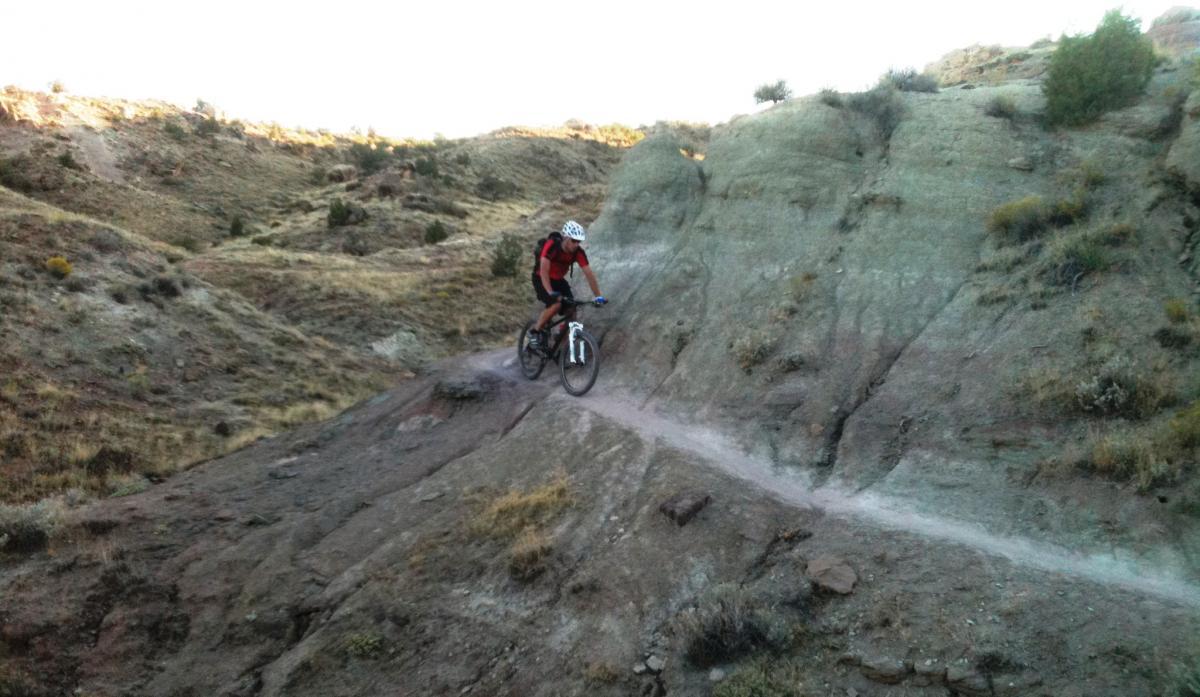 A mountain biker riding down a rocky trail surrounded by hilly terrain in natural light. The biker is wearing a helmet and riding gear, navigating a narrow path along the hillside. Sparse vegetation and rock formations are visible in the background, with varying shades of brown and green from the landscape. Lunch Loops mountain bike trail.