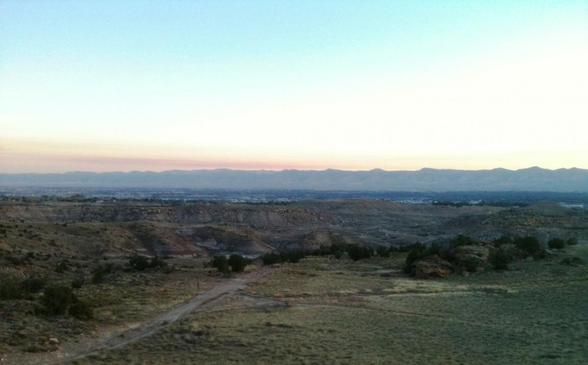 A panoramic view of a rugged landscape featuring rolling hills and mountains in the background, illuminated by the soft colors of dawn or dusk. The foreground includes sparse vegetation and a dirt path winding through the terrain, with a clear sky transitioning from light blue to softer pastel hues. Lunch Loops mountain bike trail.