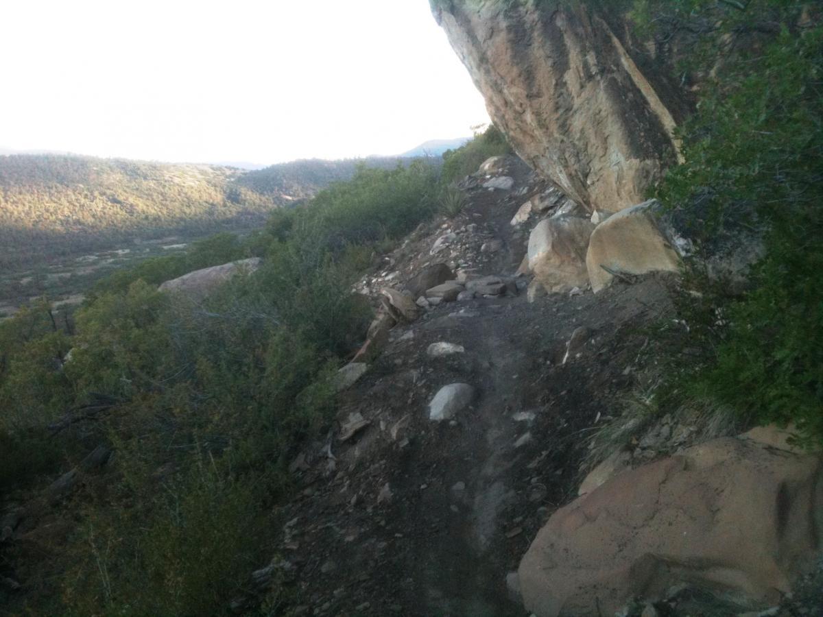 A narrow dirt trail winding along a rocky hillside, surrounded by shrubs and trees, with a distant view of rolling hills and valleys illuminated by soft morning light. Horse Gulch mountain bike trail.