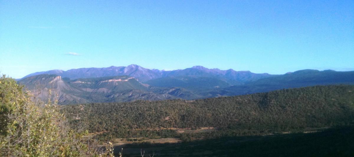 A panoramic view of rolling hills and mountains under a clear blue sky, featuring lush greenery in the foreground and distant peaks with varying elevations. The landscape showcases the natural beauty of a mountainous region. Horse Gulch mountain bike trail.