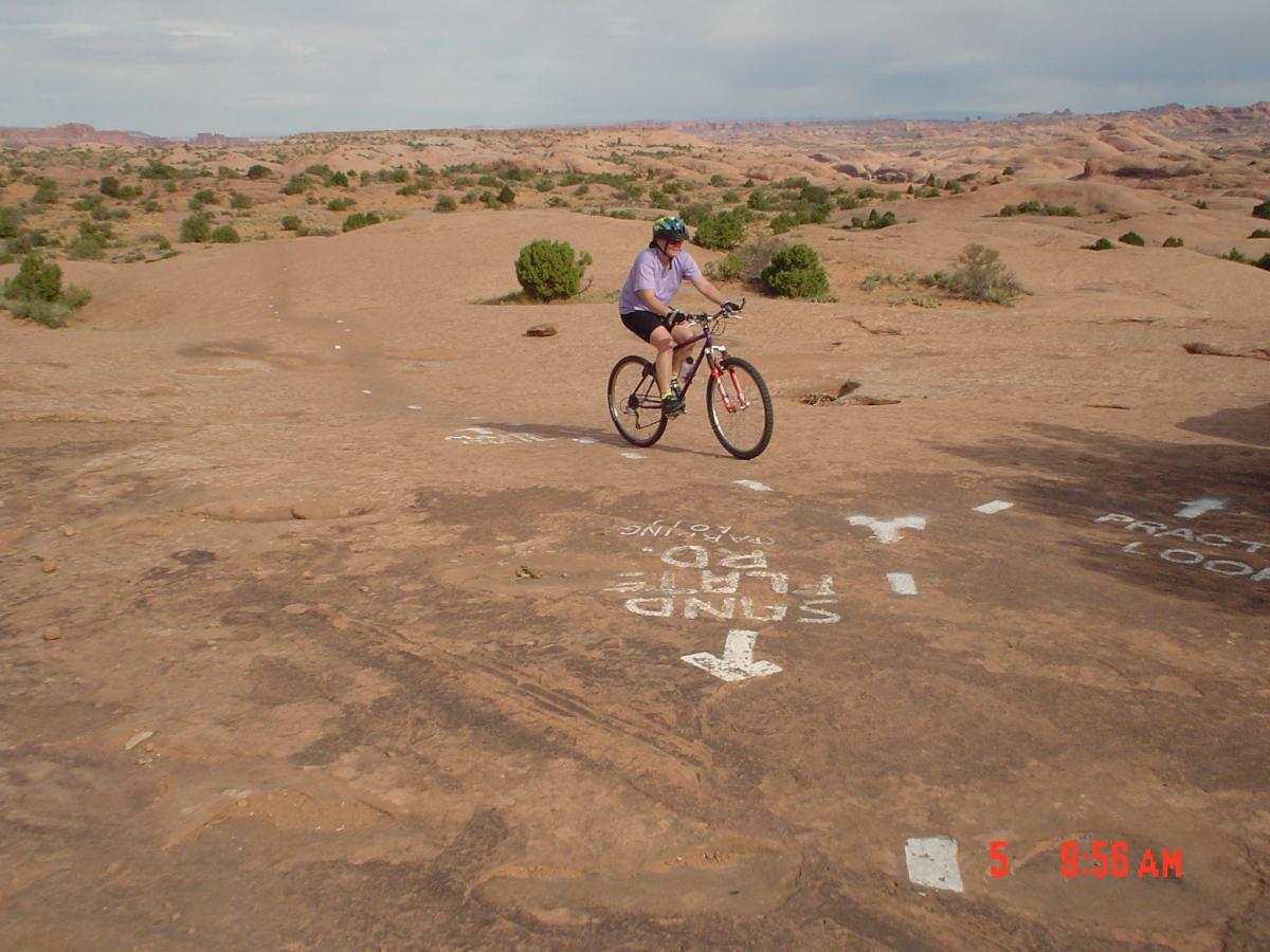 A person riding a mountain bike on a rocky trail, surrounded by a desert landscape with shrubs and rock formations. There are directional signs painted on the ground indicating different bike routes. The sky is partly cloudy, suggesting a sunny day. Slickrock mountain bike trail.