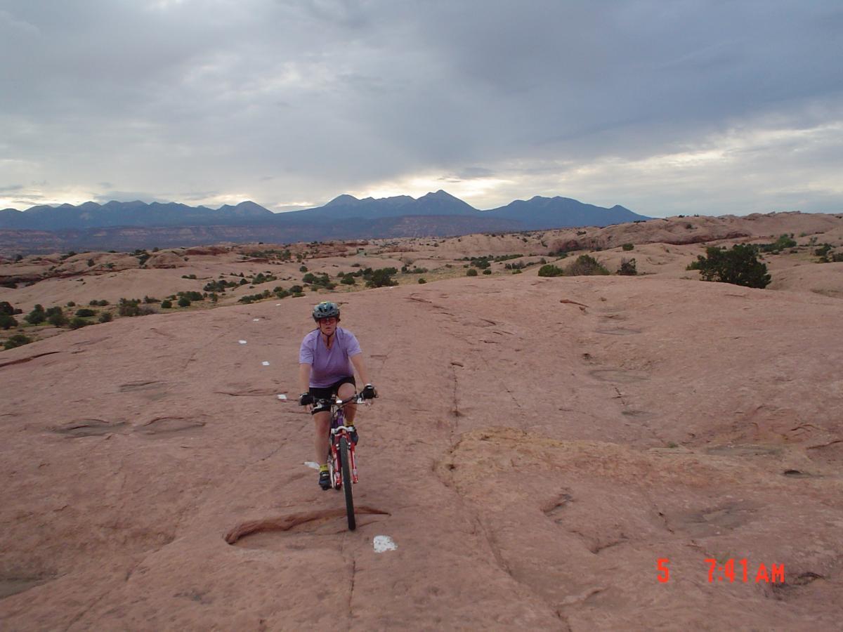 A cyclist riding a mountain bike on a rocky terrain, with expansive desert landscape and mountains in the background under a cloudy sky. Slickrock mountain bike trail.