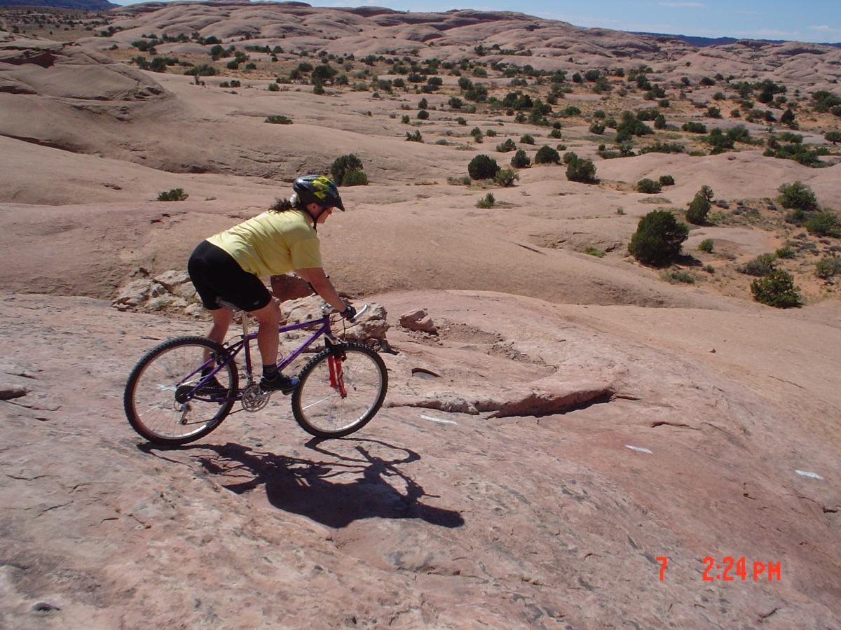 Mountain biker descending a rocky terrain in a desert landscape, wearing a yellow shirt and helmet. The scene captures the natural beauty of the area, with sparse vegetation and smooth rock formations under a clear blue sky. Slickrock mountain bike trail.