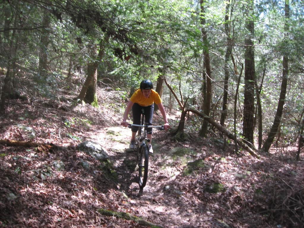 A person riding a mountain bike on a rocky trail through a forested area, surrounded by trees and underbrush. The cyclist is wearing a yellow shirt and a helmet, leaning forward as they navigate the uneven path. Sunlight filters through the foliage, creating dappled light on the ground. River Loop mountain bike trail.