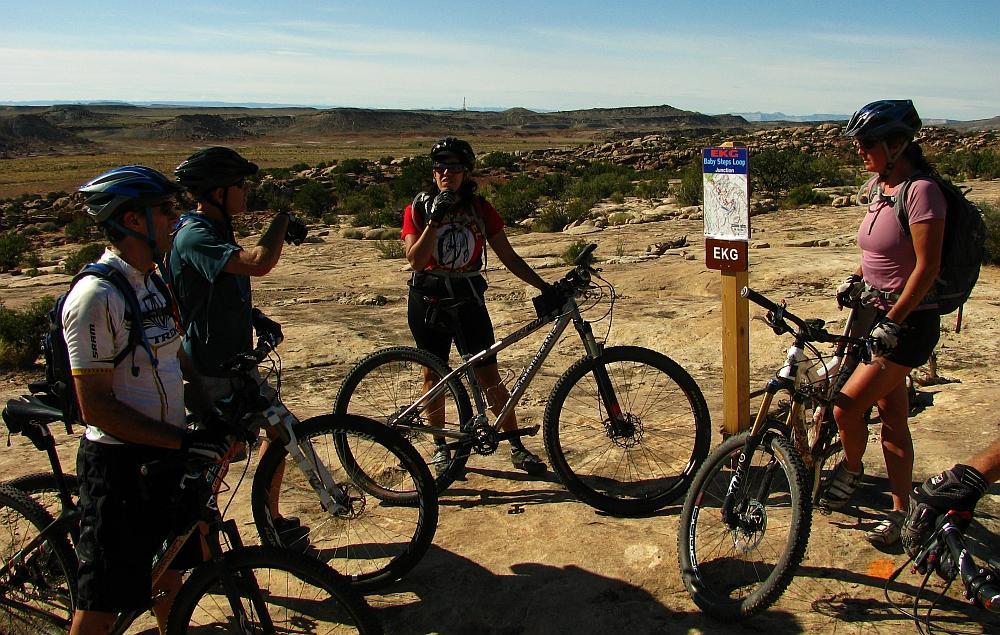A group of four mountain bikers, wearing helmets and athletic gear, gathered at a trail intersection in a rocky landscape. They are engaged in discussion, with one person gesturing towards a sign that details the "Baby Steps Loop" trail. The background features a panoramic view of rolling hills and clear blue skies. Klondike Bluffs mountain bike trail.
