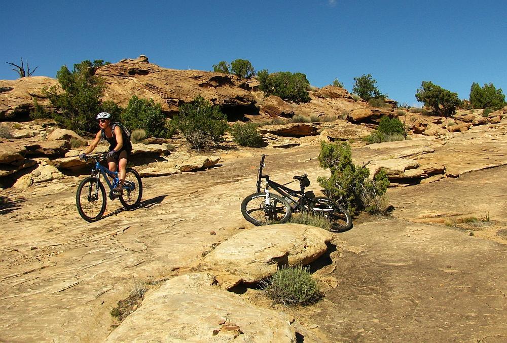 A mountain biker rides on a rocky terrain under a clear blue sky, with scrubby greenery in the background. Another bike lies propped against a rock nearby, emphasizing the outdoor adventure setting. Klondike Bluffs mountain bike trail.