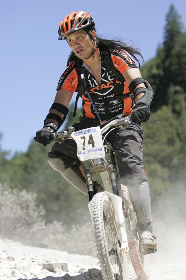 A mountain biker with long hair rides down a dusty trail during a race, wearing a vibrant orange and black jersey, knee pads, and a helmet. The cyclist