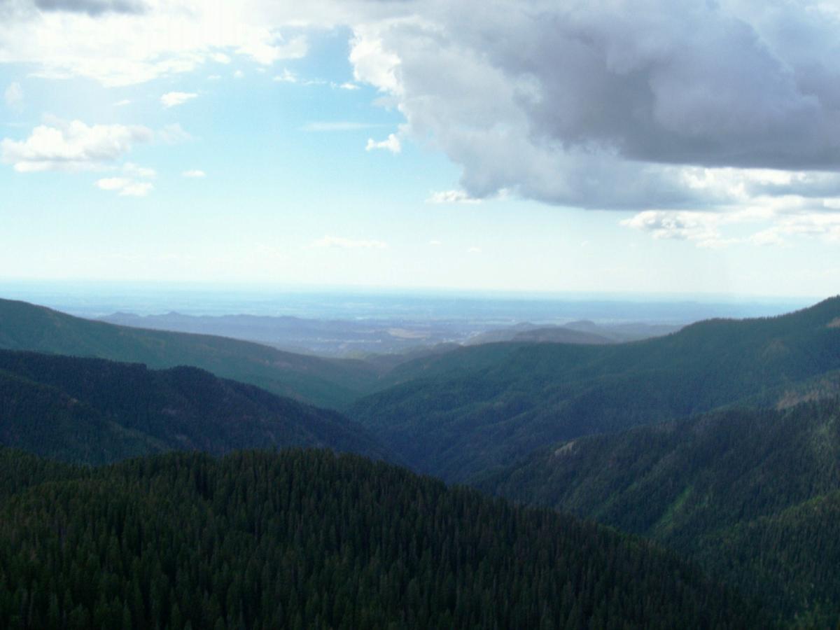 A panoramic view of rolling green mountain ranges under a cloudy sky, with distant hills fading into the horizon. The foreground features dense forests, while the background transitions to a lighter sky, suggesting a clear day. Colorado Trail: Kennebec Pass To Junction Creek mountain bike trail.