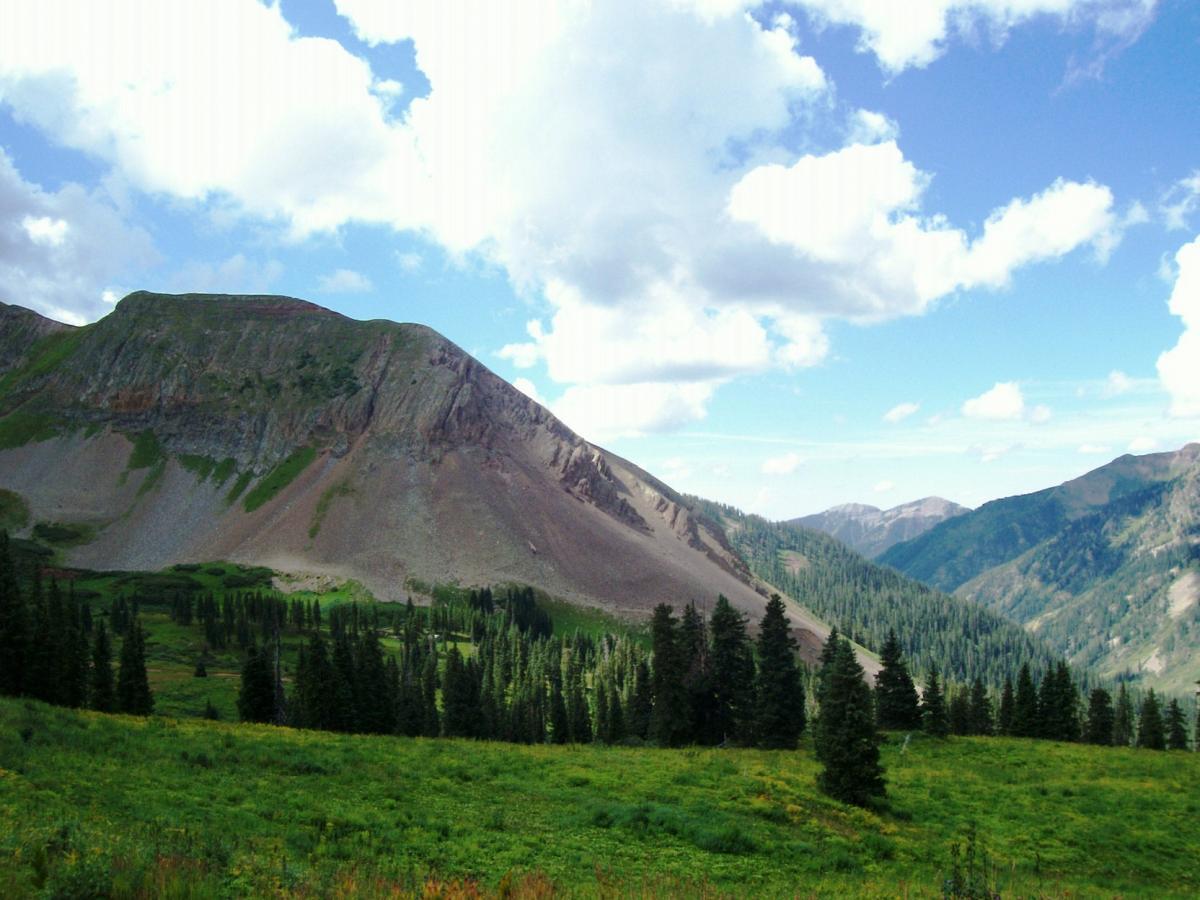 A scenic mountain landscape featuring rolling hills, rocky peaks, and a lush green valley with tall evergreen trees under a partly cloudy sky. Colorado Trail: Kennebec Pass To Junction Creek mountain bike trail.