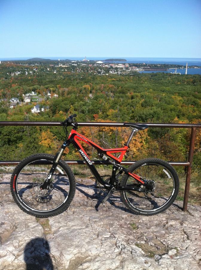 A mountain bike in vibrant red and black rests on a rocky outcrop, with a scenic view of a colorful autumn landscape and a distant body of water visible in the background. The horizon is dotted with trees showcasing fall foliage, and a small town can be seen in the valley. A metal railing runs alongside the bike, enhancing the image of adventure and exploration. Noquemanon Trails Network: South Marquette Trails mountain bike trail.