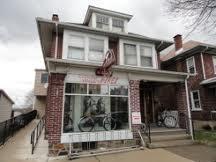 A two-story brick building with a large front window displaying bicycles inside. The building has a gabled roof and a welcoming entrance with steps leading up to it. Overcast sky in the background.