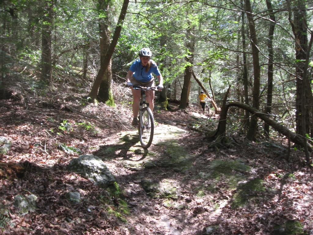A person biking on a rocky trail surrounded by lush green trees and foliage in a wooded area. Another individual is seen further along the path, contributing to an active outdoor scene. River Loop mountain bike trail.