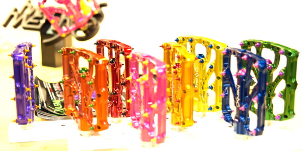 A collection of colorful bicycle pedals arranged in a row, showcasing various vibrant hues including blue, pink, yellow, green, and orange. The pedals have a metallic finish and are designed with small spikes for grip. In the background, there is a blurred logo of a bike-related brand.