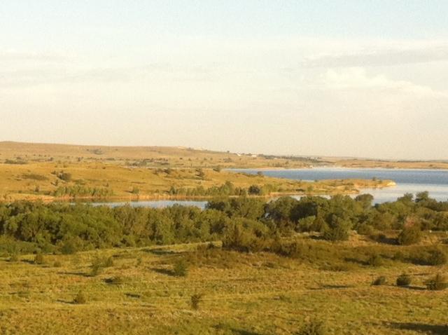 A panoramic view of a serene landscape featuring a gently flowing lake bordered by green vegetation and rolling hills under a clear sky. The scene captures the tranquil beauty of nature, showcasing a harmonious blend of water, grassland, and scattered trees. Switchgrass mountain bike trail.