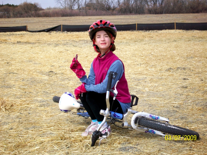 A young girl wearing a bicycle helmet and colorful gloves sits on her bike in a field. She has a playful expression and is making a peace sign with her fingers. The background features a cloudy sky and a few trees, while the ground is covered in dry grass or straw.