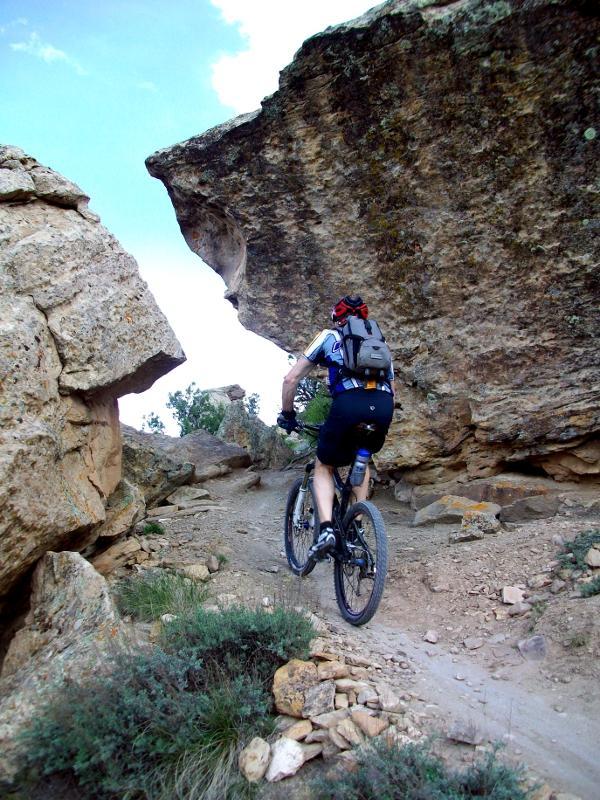 A mountain biker riding on a rocky trail surrounded by large boulders and a clear blue sky. The rider is wearing a helmet and a backpack, navigating through a rugged terrain. Moore Fun mountain bike trail.