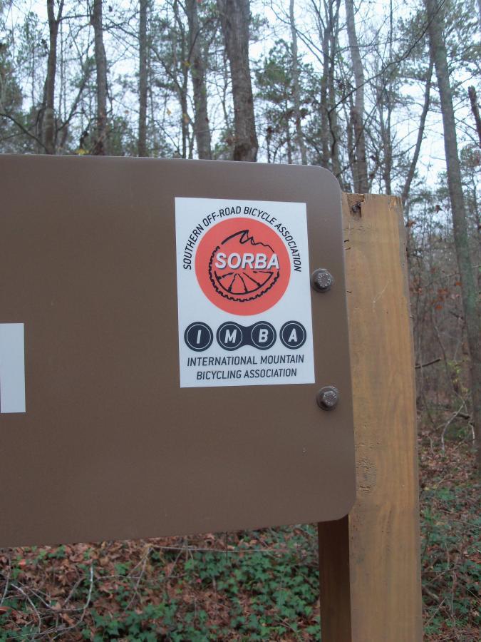 A close-up image of a trail sign featuring two logos: the Southern Off-Road Bicycle Association (SORBA) and the International Mountain Bicycling Association (IMBA). The sign is mounted on a wooden post, surrounded by trees in a wooded area. Paynes Creek mountain bike trail.