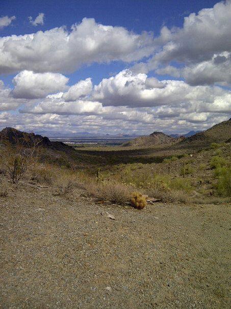 A panoramic view of a desert landscape featuring rolling hills and mountains under a partly cloudy sky. The foreground shows rocky terrain with sparse vegetation, while the background includes distant mountains and a wide valley. Trail #100 mountain bike trail.