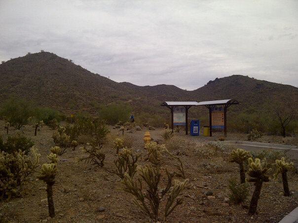 A desert landscape featuring a bus stop with a shelter and informational signage, surrounded by cacti and sparse vegetation. In the background, rugged mountains rise under a cloudy sky, creating a serene yet arid environment. Sonoran Trail mountain bike trail.