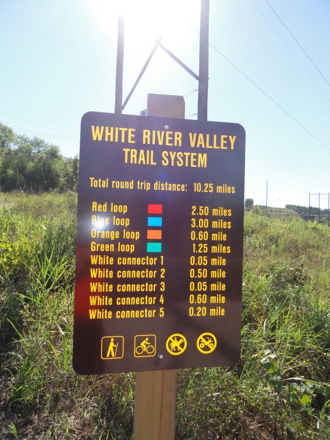 Sign indicating the White River Valley Trail System, detailing the total round trip distance of 10.25 miles and the distances of various loops and connectors: Red loop (2.50 miles), Blue loop (3.00 miles), Orange loop (0.60 miles), Green loop (1.25 miles), and five white connectors ranging from 0.05 to 0.60 miles. The sign includes icons for hiking, biking, and other trail activities. White River Valley mountain bike trail.