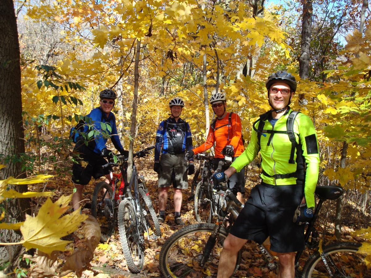 Four mountain bikers, wearing colorful cycling gear and helmets, stand beside their bikes on a wooded trail surrounded by vibrant yellow autumn leaves. The scene captures the essence of an active outdoor lifestyle in nature during fall. Frick Park mountain bike trail.