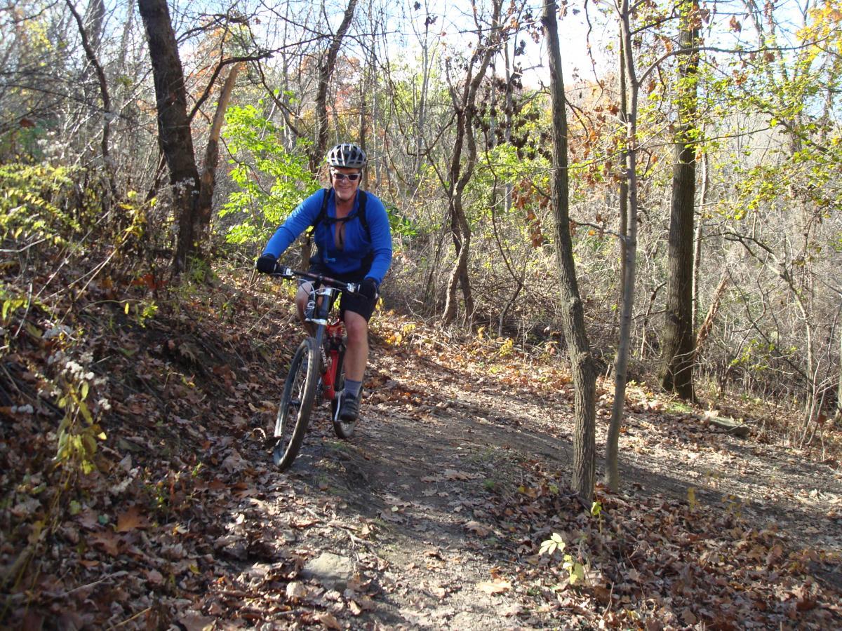 A person riding a mountain bike on a dirt trail surrounded by trees and autumn foliage. The cyclist is wearing a blue long-sleeve shirt, a helmet, and shorts, and is navigating the winding path through the forest. Leaves are scattered on the ground, indicating the fall season. Frick Park mountain bike trail.
