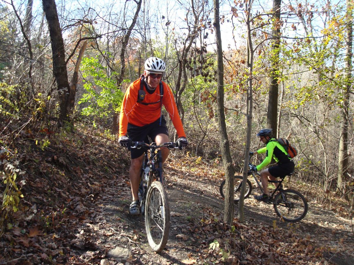 Two mountain bikers navigate a dirt trail surrounded by trees in an autumn setting. One rider wears an orange long-sleeve shirt and is slightly ahead, while the other, in a bright green shirt, follows closely behind. The ground is covered with fallen leaves, and the atmosphere is lively with greenery in the background. Frick Park mountain bike trail.