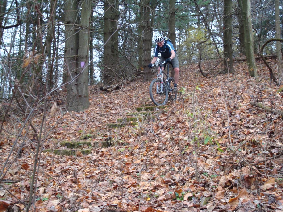 A mountain biker navigates a steep, leaf-covered hill in a wooded area, surrounded by trees. The biker is wearing a helmet and riding gear, focusing on the path ahead while descending the incline. Deer Lakes Park mountain bike trail.