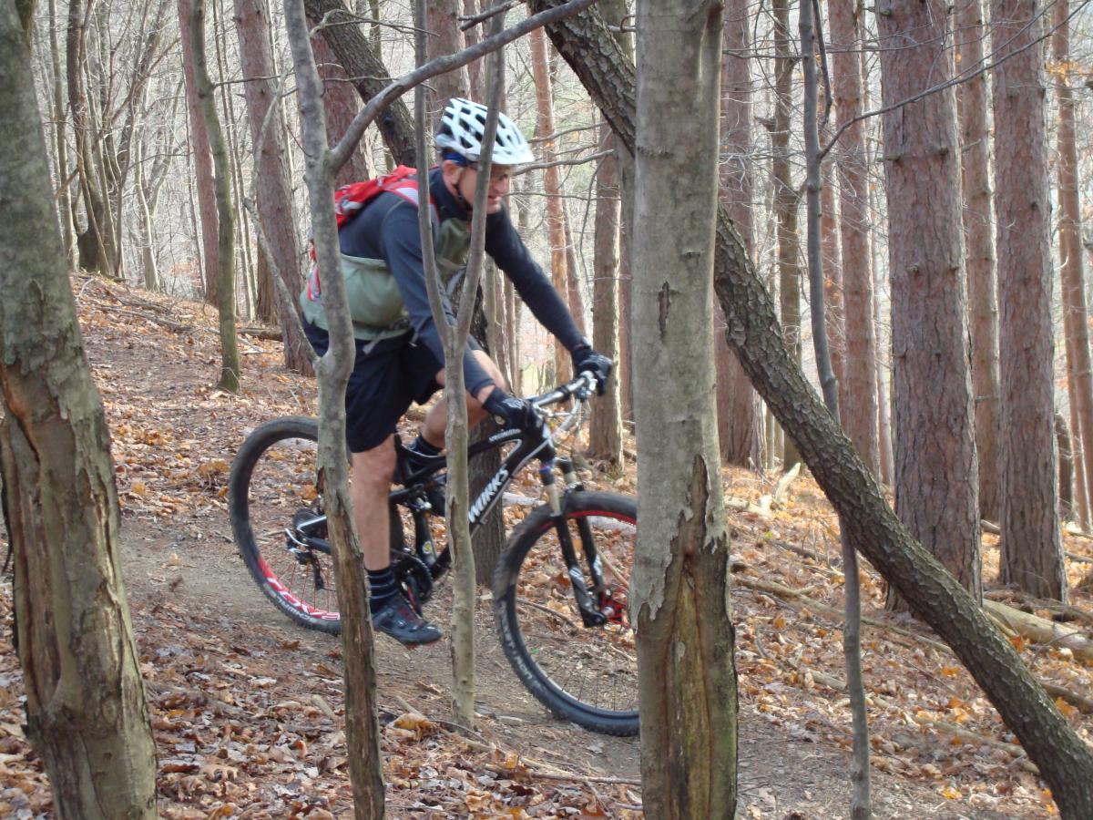 Mountain biker navigating a trail through a wooded area, surrounded by trees and autumn leaves on the ground. The rider is wearing a helmet and a backpack, focused on the path ahead. North Park mountain bike trail.