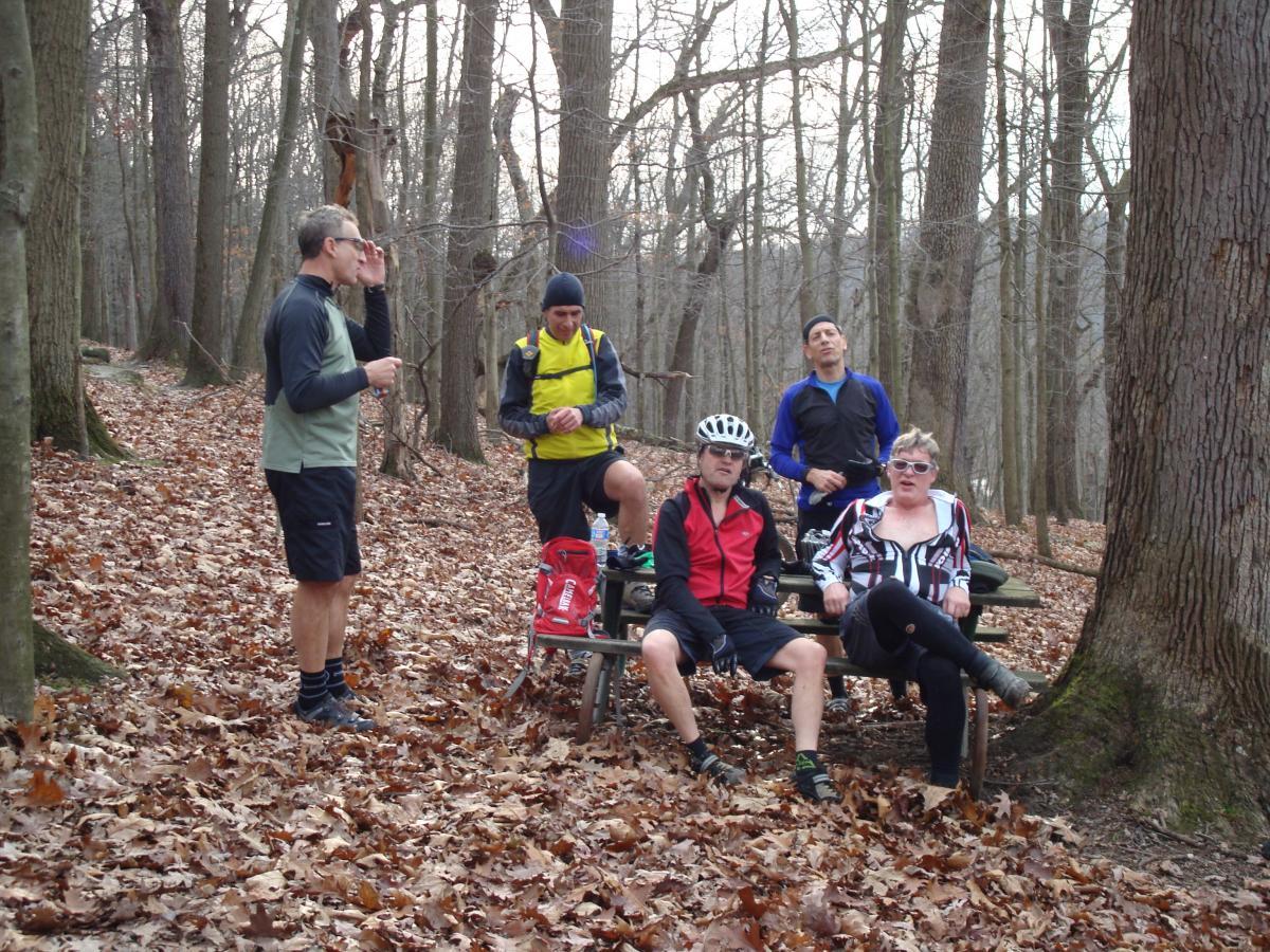 A group of five people taking a break in a wooded area during a cycling or hiking trip. They are gathered around a picnic table covered in fallen leaves, with trees visible in the background. One person is standing and drinking from a bottle, while others are seated, some in cycling attire. The scene captures a casual, outdoor atmosphere. North Park mountain bike trail.