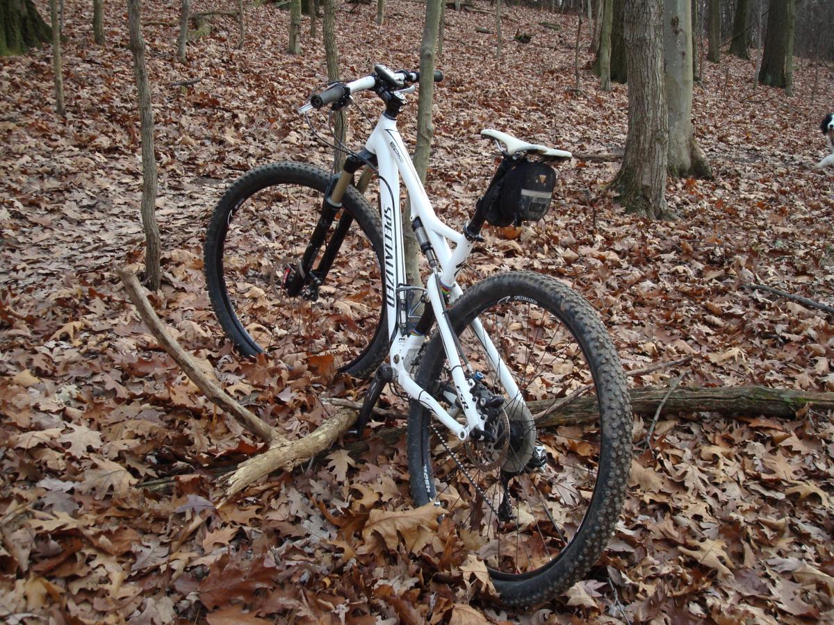 A mountain bike stands on a bed of fallen leaves in a wooded area, surrounded by trees. The bike features a white frame and black wheels, with a small bag mounted on the rear. The ground is covered with leaves and twigs, indicating a natural outdoor setting. North Park mountain bike trail.