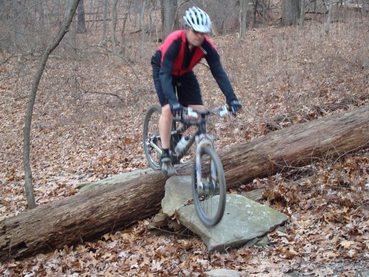 A mountain biker navigating over a large rock while balancing on a log in a forested area covered with fallen leaves. North Park mountain bike trail.