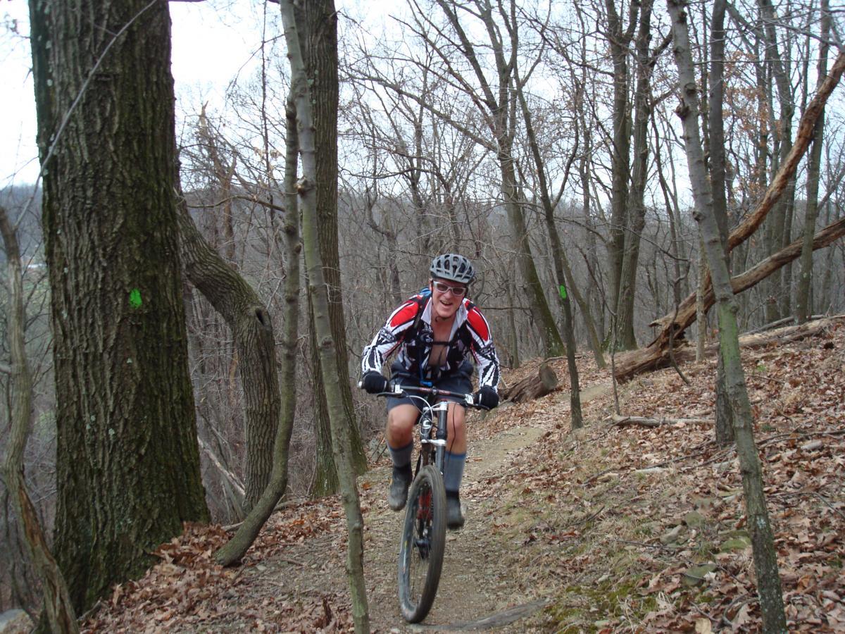 A mountain biker riding a trail through a wooded area, with bare trees and autumn leaves on the ground. The cyclist is wearing a helmet and athletic clothing, smiling as they navigate the uneven terrain. Hartwood Acres mountain bike trail.
