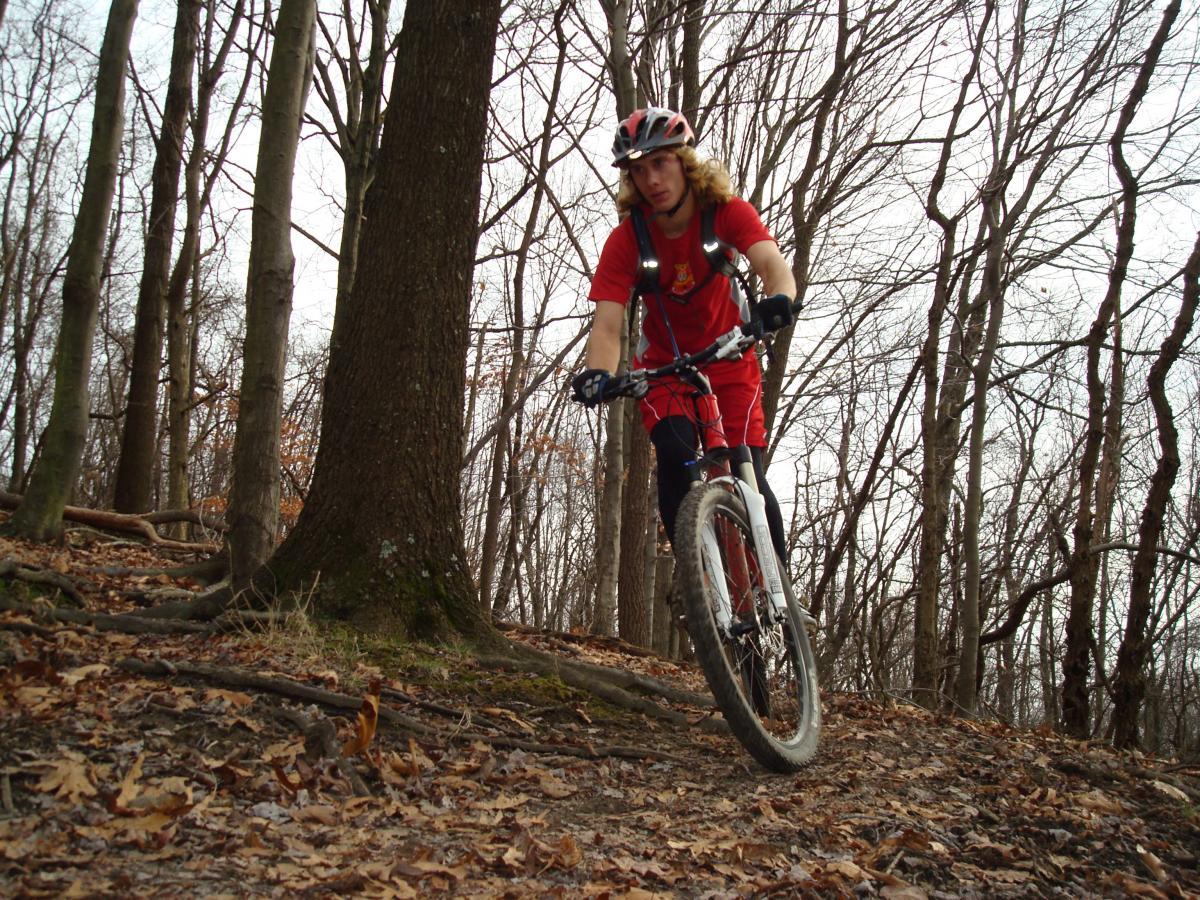 A mountain biker in a red outfit navigates a winding trail surrounded by bare trees and fallen leaves. The cyclist is focused, leaning slightly forward on the bike as they ride along a forest path. Hartwood Acres mountain bike trail.