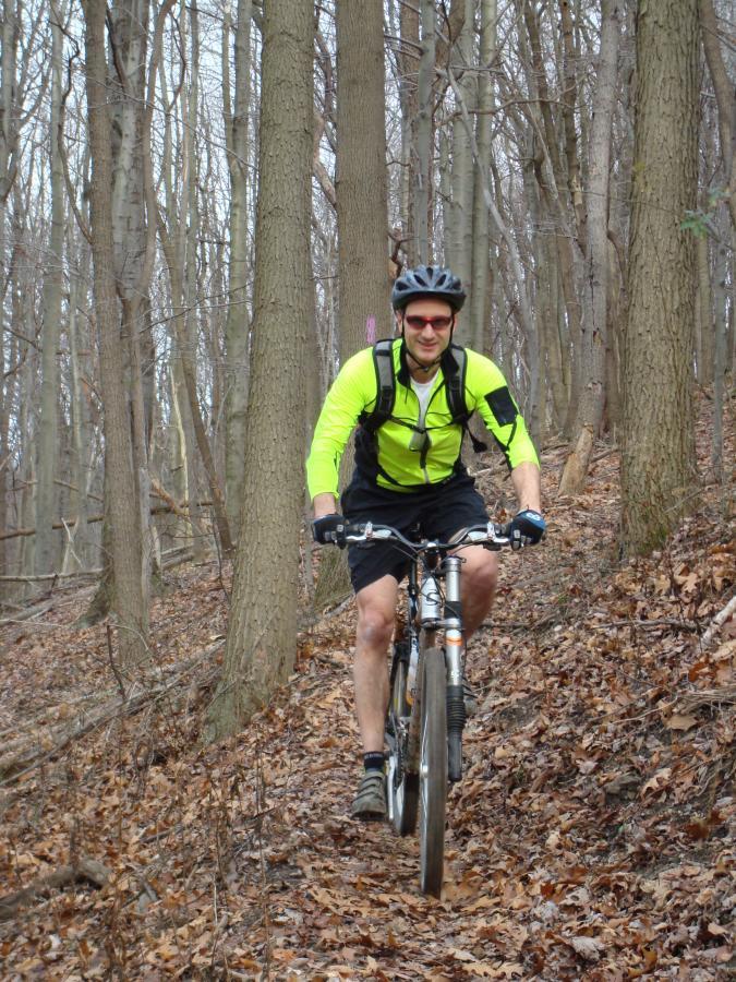 A mountain biker wearing a bright yellow and black cycling outfit, along with a helmet and sunglasses, rides through a trail surrounded by bare trees and fallen leaves in a forested area. Hartwood Acres mountain bike trail.