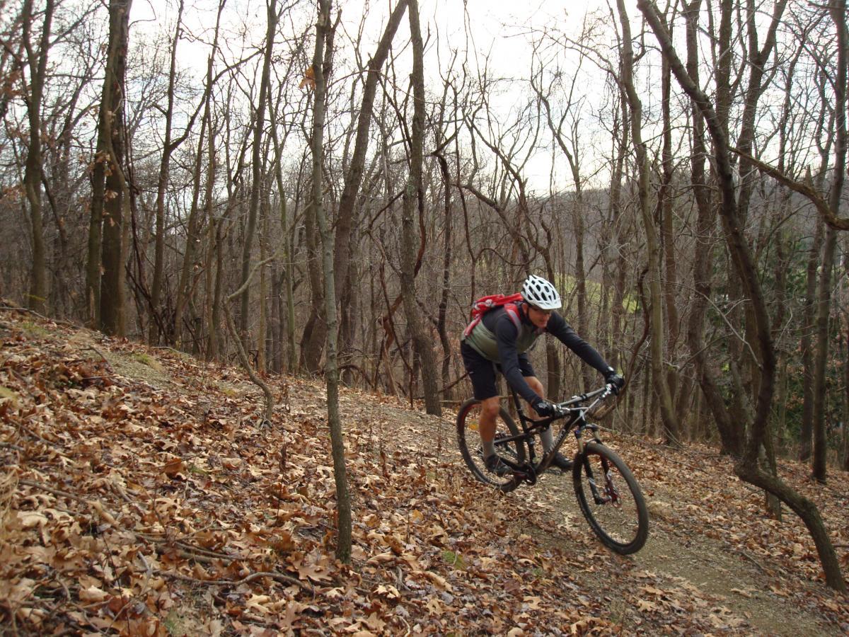 A mountain biker navigating a leaf-covered trail through a wooded area with bare trees in the background. The cyclist wears a helmet and a backpack, leaning into the turn on the bike. Hartwood Acres mountain bike trail.