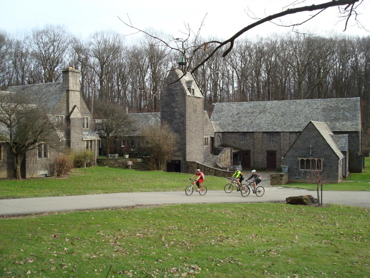 Three cyclists riding on a paved path in front of a large stone building complex surrounded by grass and bare trees. The buildings feature sloped roofs and a mix of architectural styles, with one structure appearing to have a tower. The scene shows a tranquil outdoor setting. Hartwood Acres mountain bike trail.