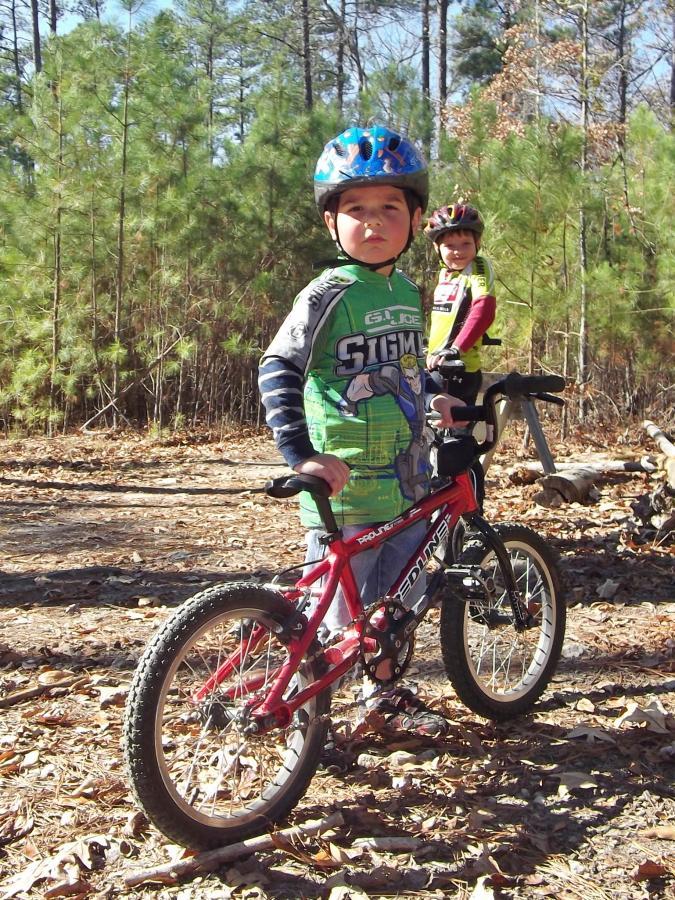 Two children stand on a dirt path in a wooded area, each with a bicycle. The child in the foreground wears a colorful green helmet and a matching outfit featuring a superhero design. They strike a confident pose beside their red bicycle. The second child, in the background, wears a yellow top and a black helmet, smiling and holding a bike. Sunlight filters through the trees, illuminating the scene. Harbison State Forest mountain bike trail.