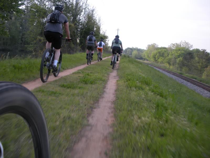 A group of five mountain bikers riding along a dirt trail next to a railway track, surrounded by greenery. The perspective shows the rear view of the cyclists as they navigate the path. Augusta Canal mountain bike trail.