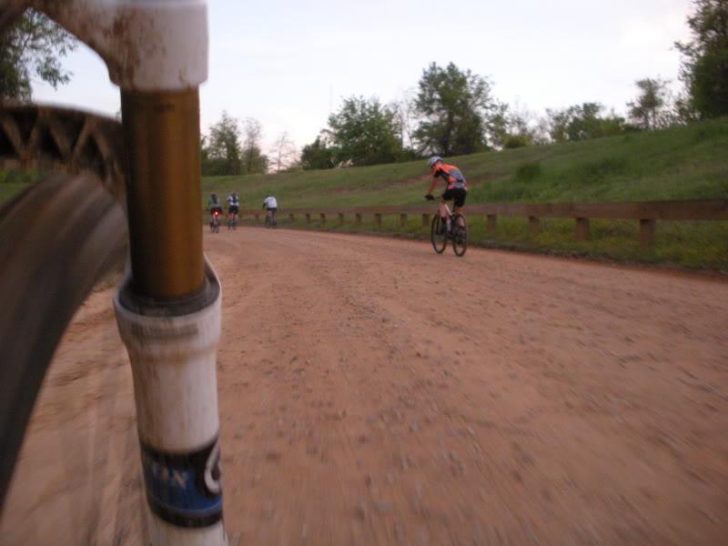 A low-angle view of a mountain biking trail, capturing the front wheel of a bike in the foreground and several cyclists riding in the background. The trail is unpaved with a sandy texture, surrounded by green grass and trees under a twilight sky. Augusta Canal mountain bike trail.