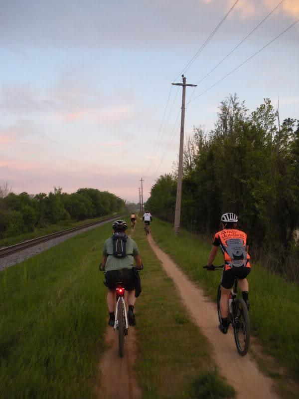 A group of cyclists riding along a dirt path adjacent to a railway track, surrounded by greenery and trees. The sky is tinged with soft colors as the sun sets, creating a tranquil outdoor scene. One cyclist is wearing an orange shirt, while others are dressed in casual biking attire. Augusta Canal mountain bike trail.
