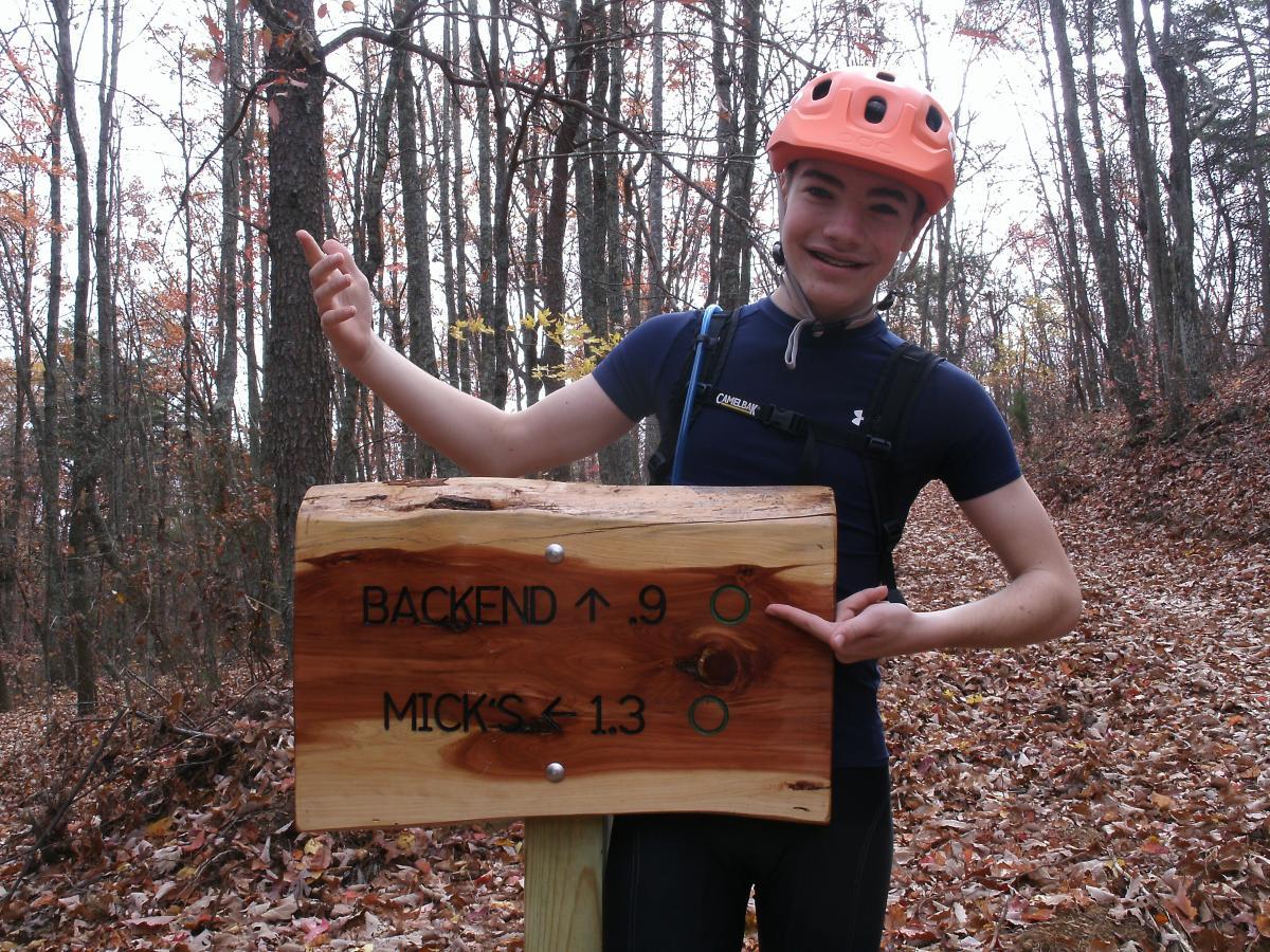 A young person wearing a bright orange helmet and athletic clothing stands next to a wooden trail sign in a forest. The sign indicates two mountain biking trails, with directional arrows pointing to "BACKEND" with a distance of 0.9 miles and "MICK'S" with a distance of 1.3 miles. The surroundings feature trees with autumn foliage and scattered leaves on the forest floor. Five Points mountain bike trail.