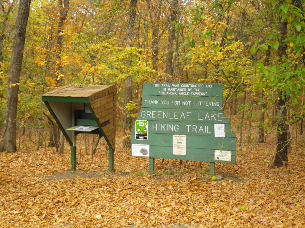 Signage at the entrance to the Greenleaf Lake Hiking Trail, with a wooden structure for trail information and a "Thank you for not littering" message. The background features trees with autumn foliage and a ground covered in fallen leaves. Oklahoma "ankle Express" Hiking Trail - Greenleaf State Park mountain bike trail.