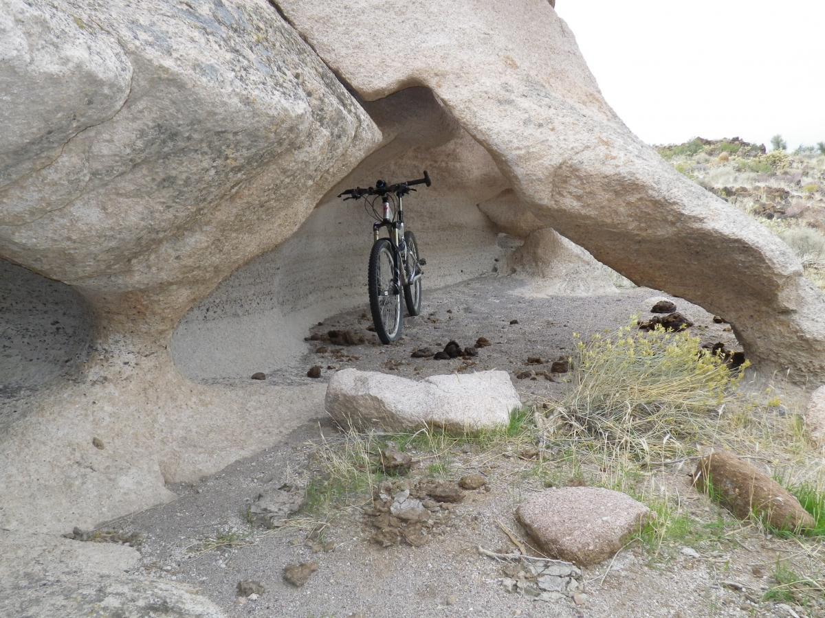 A mountain bike is parked under a large rock formation, surrounded by gravel and small boulders in a natural landscape. The area appears arid, with sparse vegetation in the background. The rock structure creates a sheltered space for the bike. Foothills Rim Trail mountain bike trail.