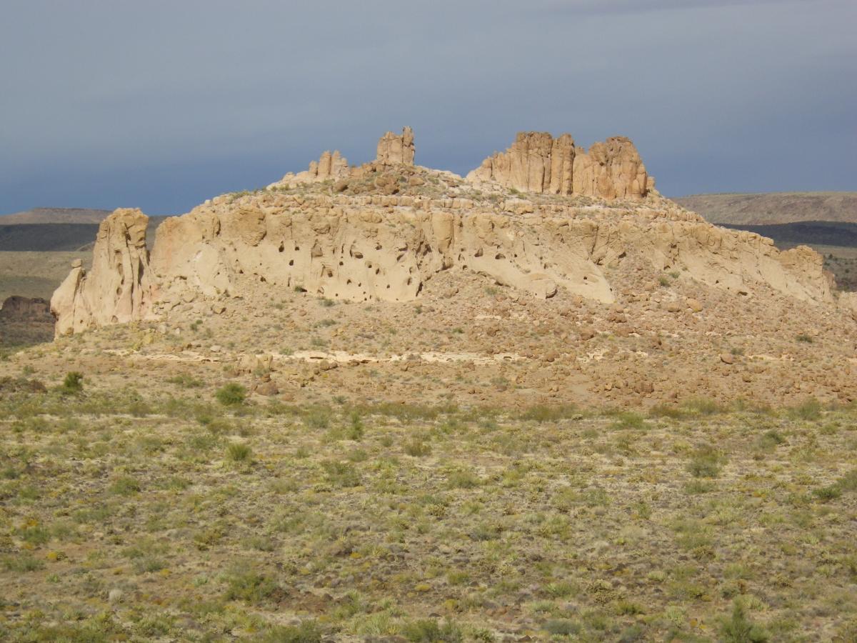 A rocky mesa formation with layered cliffs and eroded features, surrounded by a sparse landscape of low vegetation and shrubs under a cloudy sky. Foothills Rim Trail mountain bike trail.