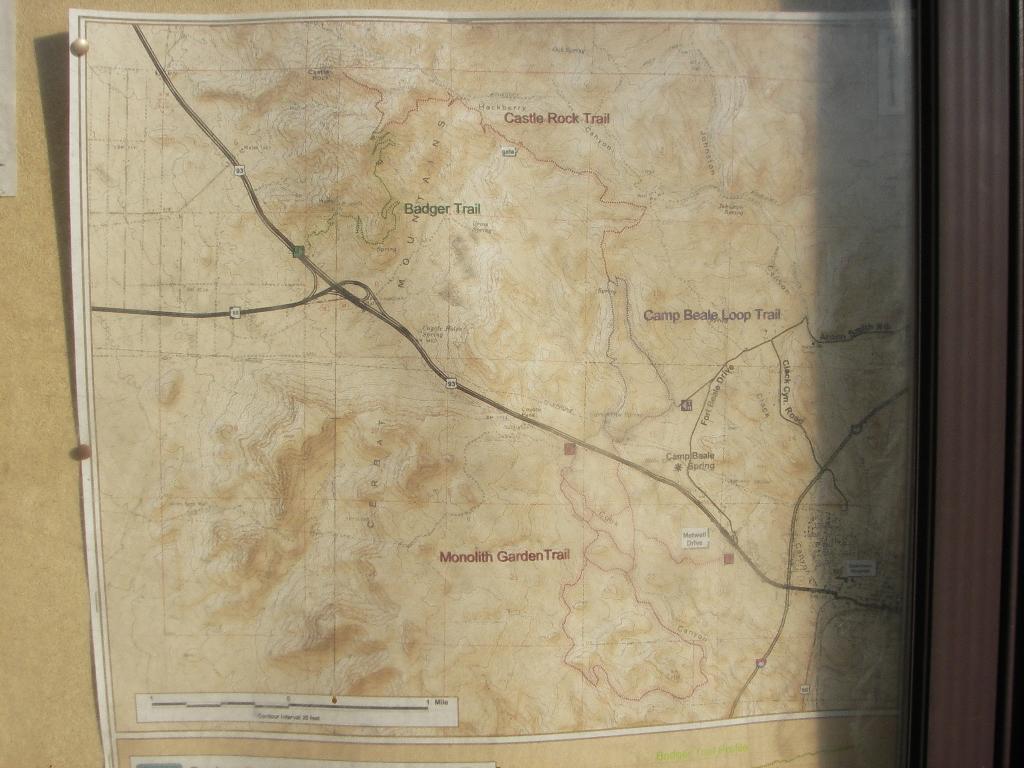 A detailed map showing various trails, including Badger Trail, Castle Rock Trail, Camp Beale Loop Trail, and Monolith Garden Trail, with terrain features and roadways indicated. The map appears weathered and is mounted on a display board. Cerbat Foothills Recreation Area mountain bike trail.