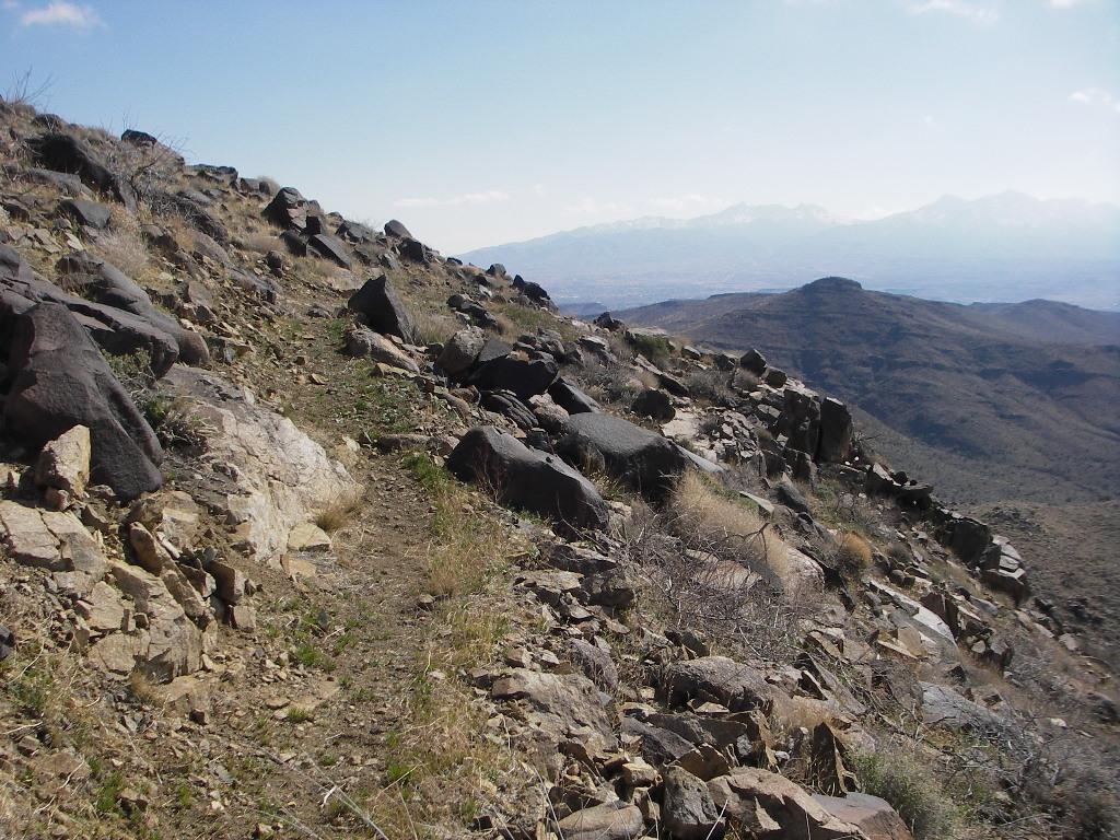 A rugged mountain trail winding through rocky terrain, with sparse vegetation and distant mountain ranges under a clear blue sky. The path is bordered by large boulders and dry grass, leading towards a scenic valley. Cerbat Foothills Recreation Area mountain bike trail.