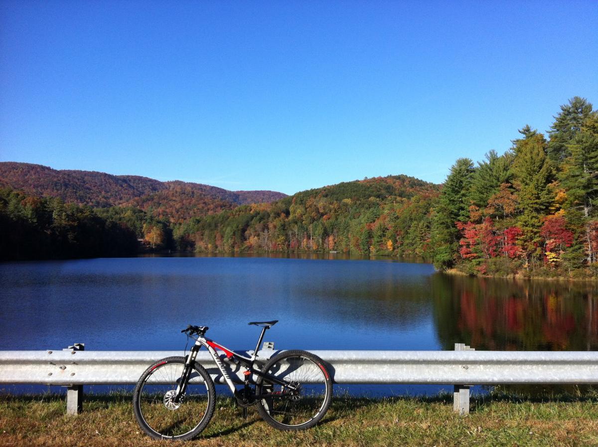A mountain bike rests against a guardrail beside a calm lake, surrounded by vibrant autumn foliage on the hills in the background. The clear blue sky reflects in the water, creating a serene and picturesque outdoor scene. Unicoi State Park mountain bike trail.