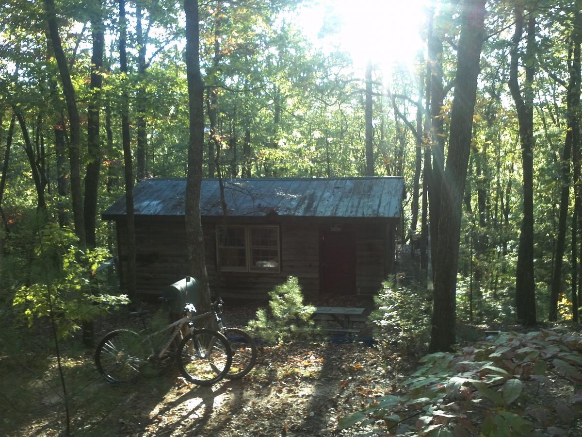 A rustic wooden cabin surrounded by lush green trees, with sunlight streaming through the foliage. A bicycle leans against a tree in the foreground, and a picnic table is visible near the cabin entrance. The scene conveys a tranquil, natural setting, ideal for outdoor relaxation. Bull / Jake Mountain mountain bike trail.