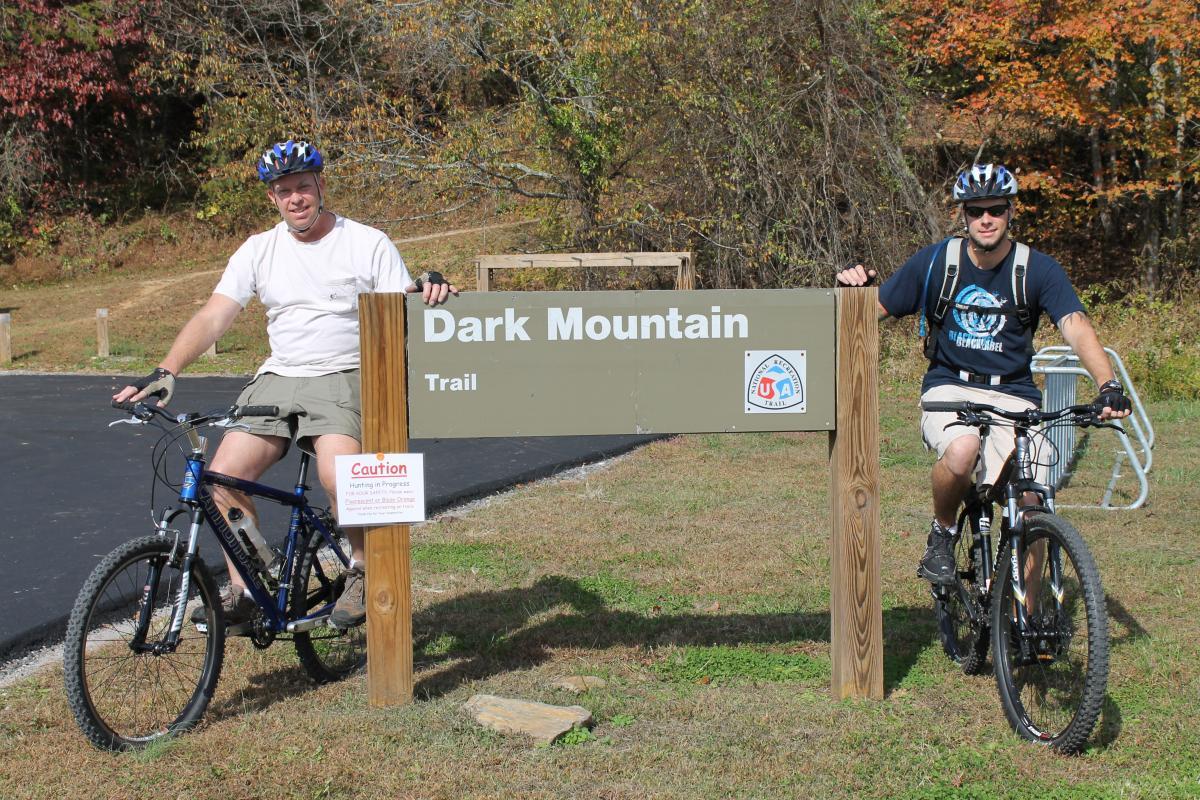 Two men on mountain bikes pose next to a sign for the Dark Mountain Trail. The sign is brown with white lettering and includes a caution notice about hunting in progress. The background features fall foliage with colorful leaves. Dark Mountain Trail mountain bike trail.